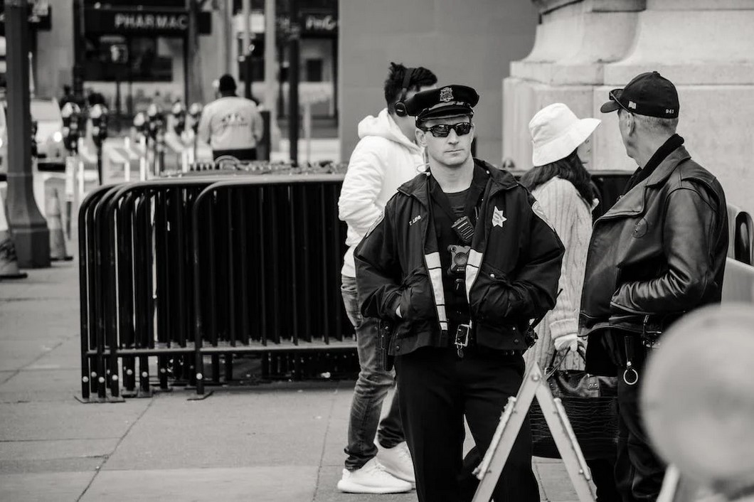 Ein Polizist in Uniform mit Sonnenbrille steht im Stadtgebiet und unterhält sich mit einem anderen Mann in Lederjacke.
